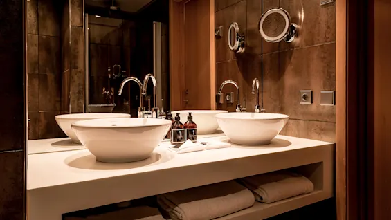 Two white sinks on a counter with a mirror and toiletries, in a warmly lit bathroom with dark tiles.