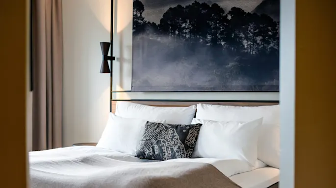 A neatly made bed with white pillows and a patterned throw pillow rests under a large painting of a misty forest. Soft lighting and curtains frame the interior.
