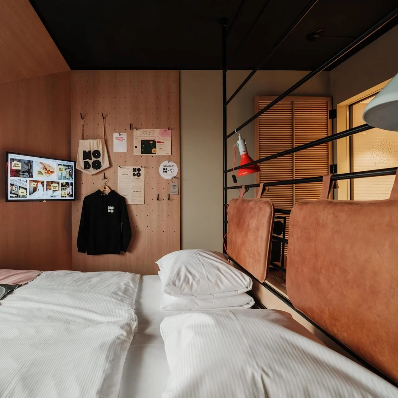 Bed with white linens sits in a cozy room. Nearby, a wall displays a black garment and pegboard with various items, including a bag labeled "NORD," under a mounted screen showing images.