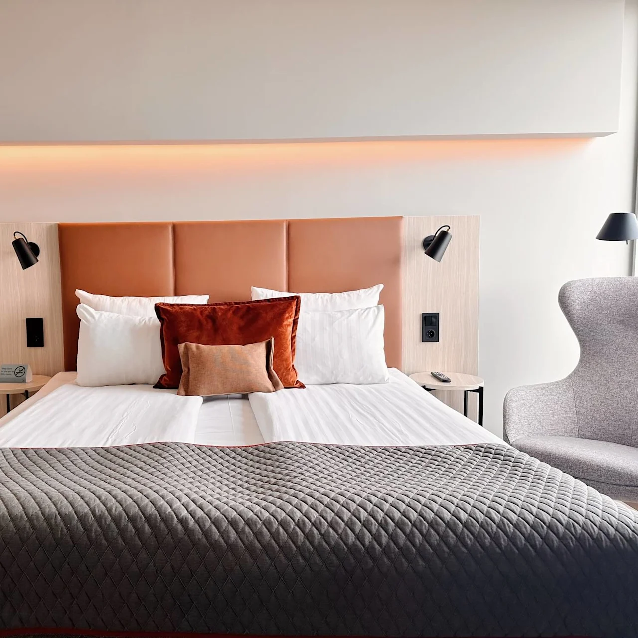 A neatly made bed with brown and white pillows rests in a modern room; a chair and lamps add to the cozy setting. Text: "No smoking."