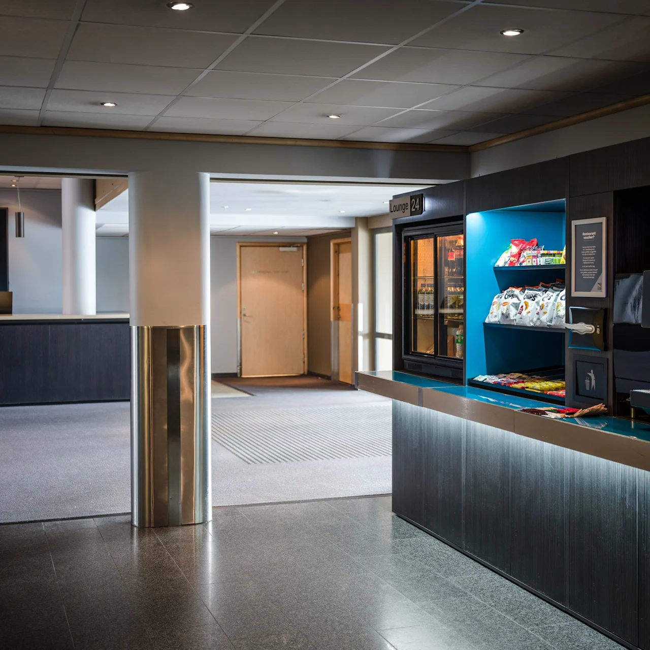 Snacksdisk som visar chips och drycker, placerad i ett modernt loungeområde. Skylt med texten "Lounge 24." En pekskärm visar "Kaffeto 20 SEK." Receptionen synlig i bakgrunden.
