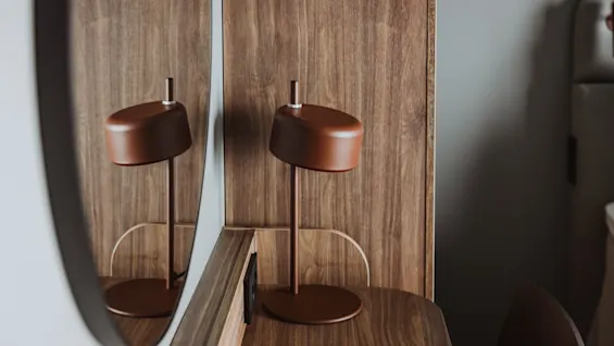 Brun skrivbordslampa på ett träbord, speglas i en närliggande spegel. En närliggande stol och grå vägg antyder en modern interiör.