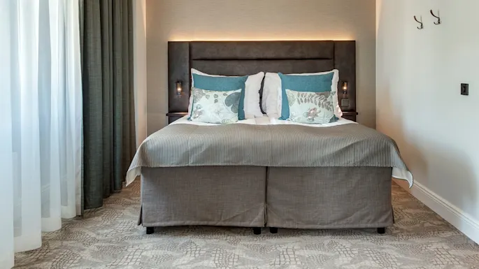 En pent oppredd seng med dekorative puter mot et polstret hodegjerde, plassert mellom to veggmonterte nattbordslamper i et godt opplyst, moderne soverom.