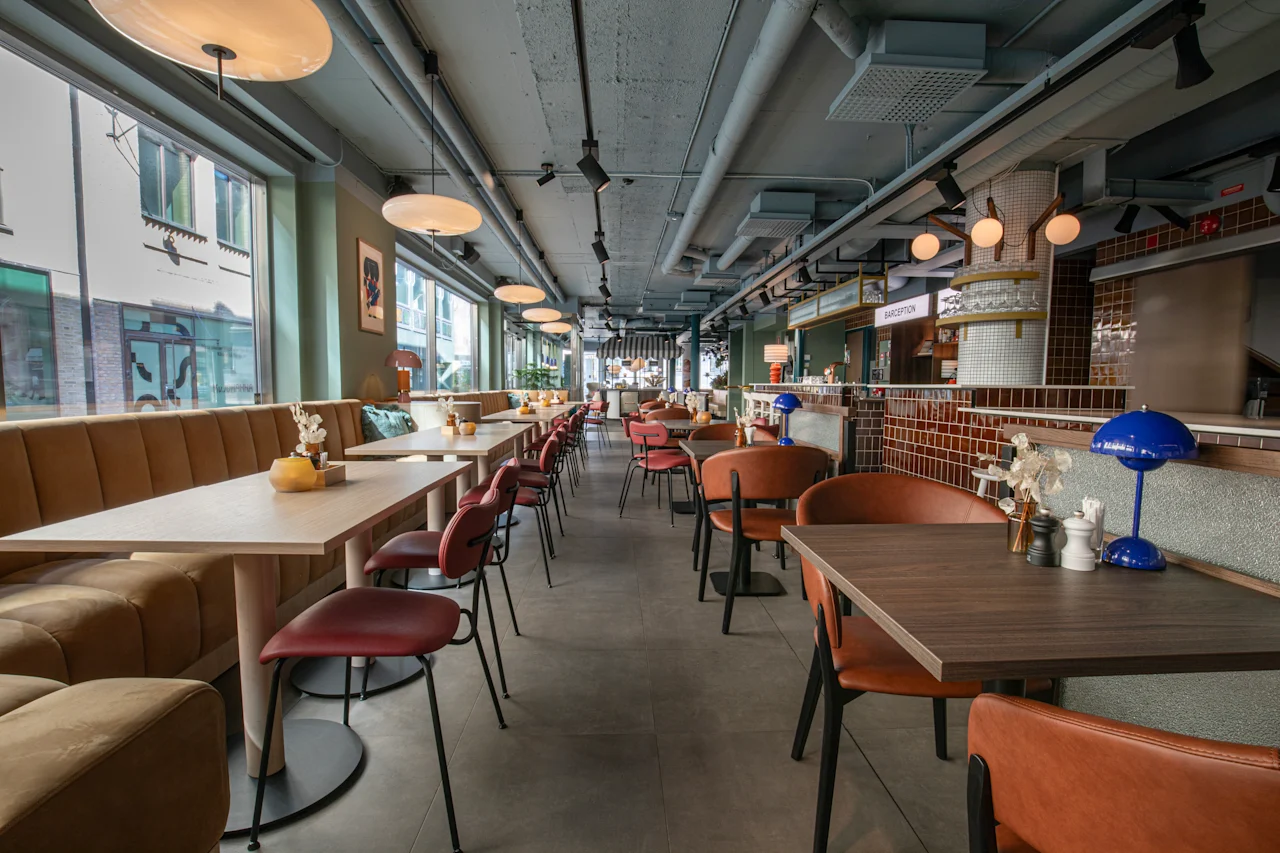 Et moderne og indbydende restaurantinteriør med borde, stole og båse langs store vinduer. En receptionsdisk er synlig.