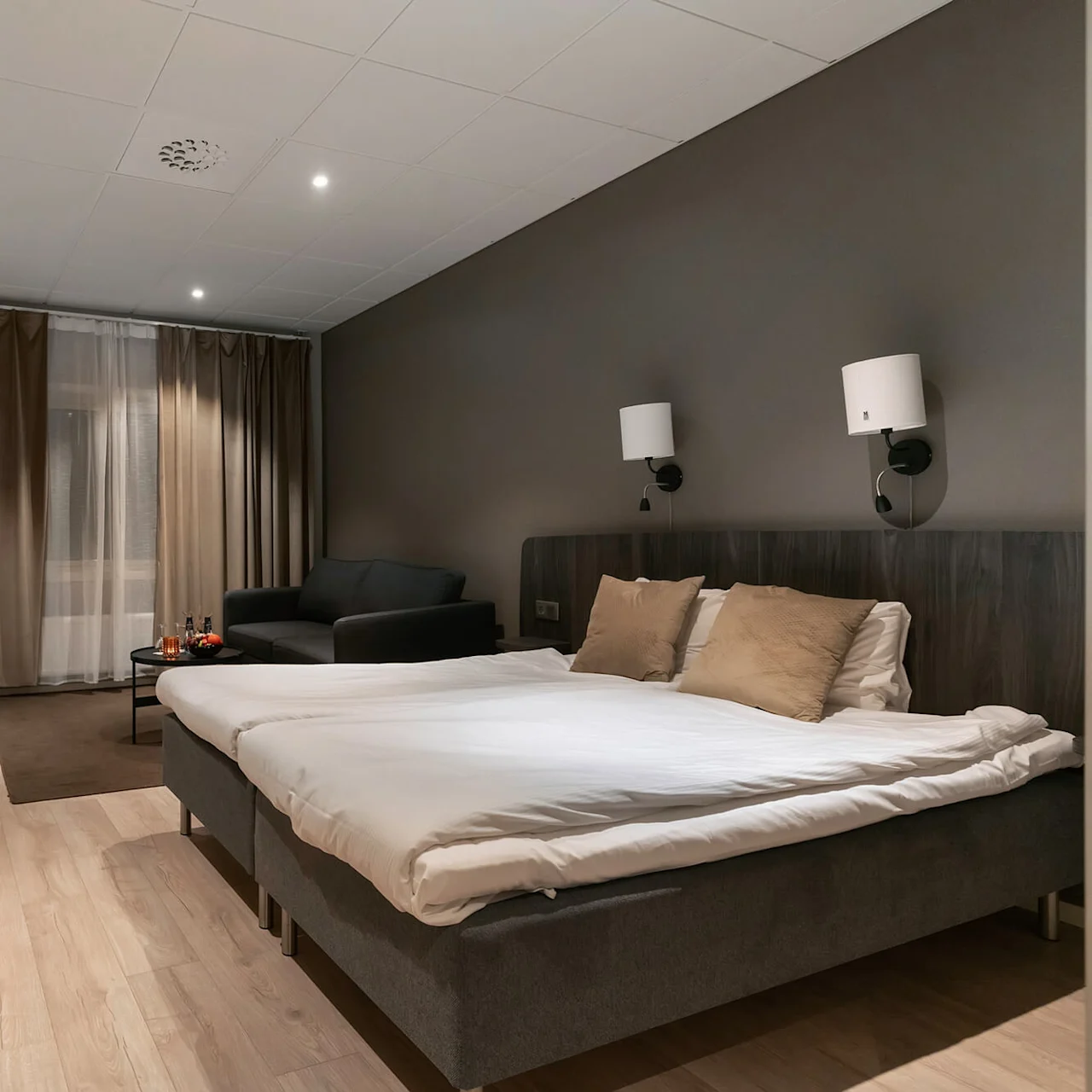 En pænt redt seng med puder står ved siden af væglamper i et moderne hotelværelse. Et skrivebord med en stol og TV er til venstre, med en sofa og gardiner bagved.
