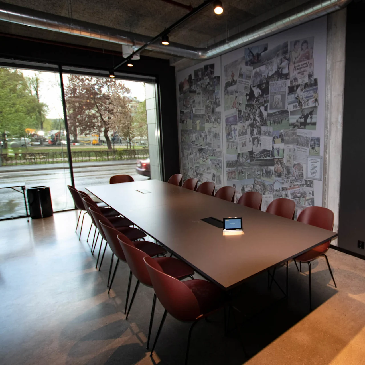 Et langt konferencetable omgivet af flere stole har en tablet, der viser "No upcoming meetings." Store vinduer afslører en gade og løv udenfor. En væg har forskellige avisudklip.