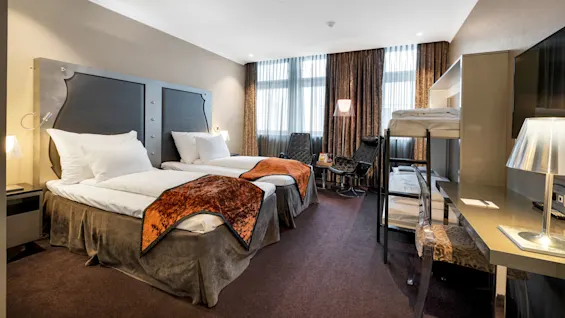 To enkeltsenge med orange tæpper er pænt arrangeret i et hotelværelse, sammen med en køjeseng, stole, skrivebord og gulvlampe mod store gardinvinduer.