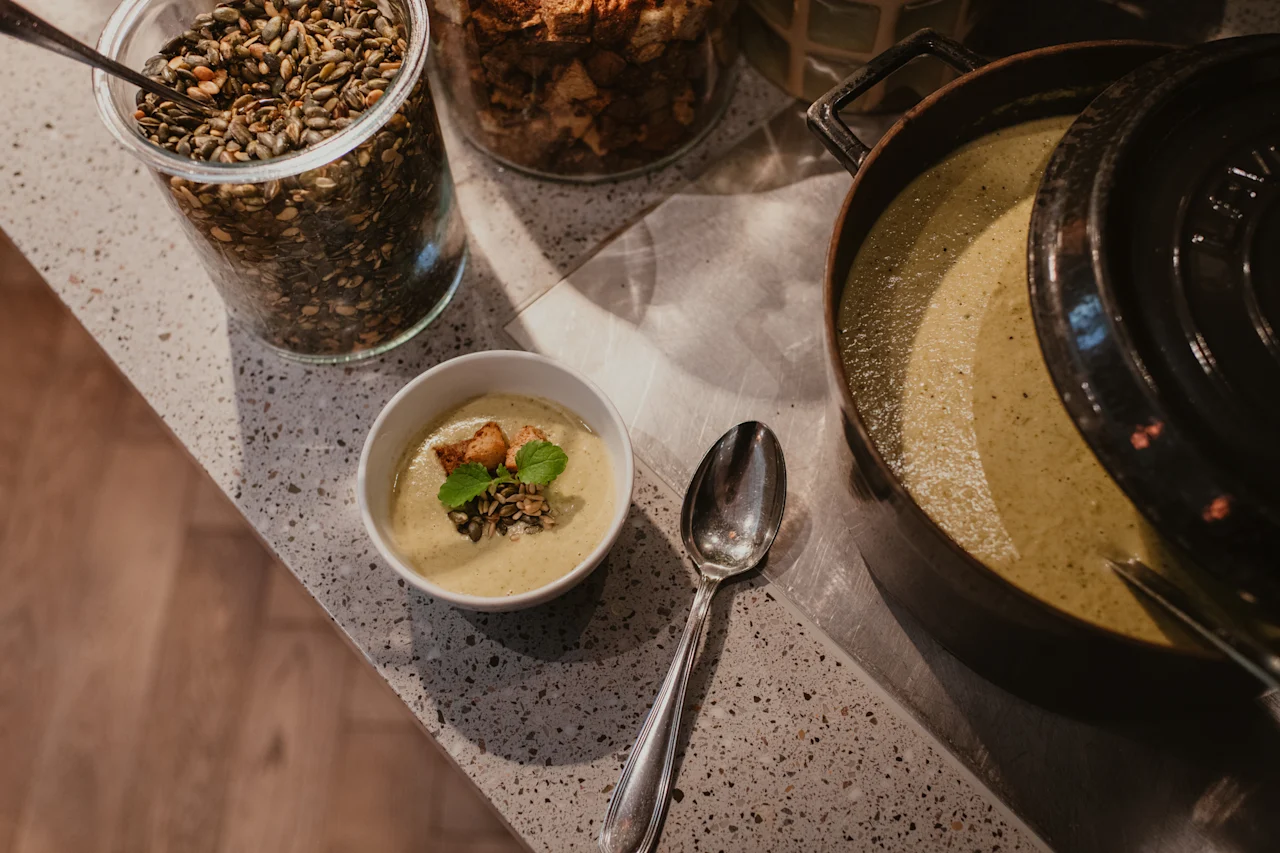 Kremet suppe i en bolle med krutonger og frø, ved siden av en stor gryte og toppingglass, som antyder et selvbetjent måltid.