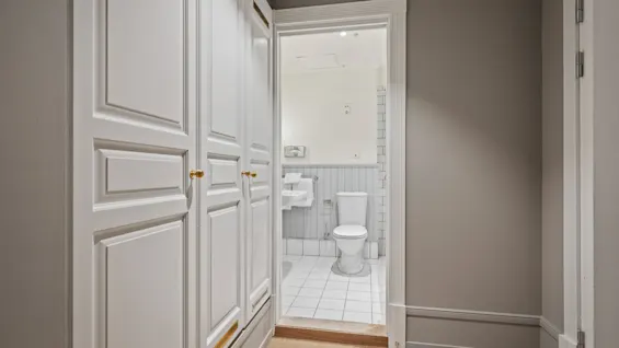 A hallway with white wardrobes on the left, leading into a bright bathroom with a toilet and sink.