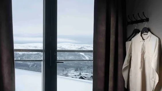 En pænt hængt hvid skjorte på en krog ved et vindue, med udsigt over et snedækket bjerglandskab, i et svagt oplyst rum med trukne gardiner og en seng.