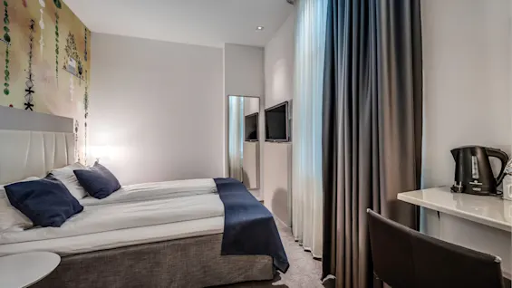 En snyggt bäddad hotellsäng med blå kuddar och filt står bredvid ett runt bord; i närheten finns en TV och gardiner i ett modernt rum. En vattenkokare står på skrivbordet.