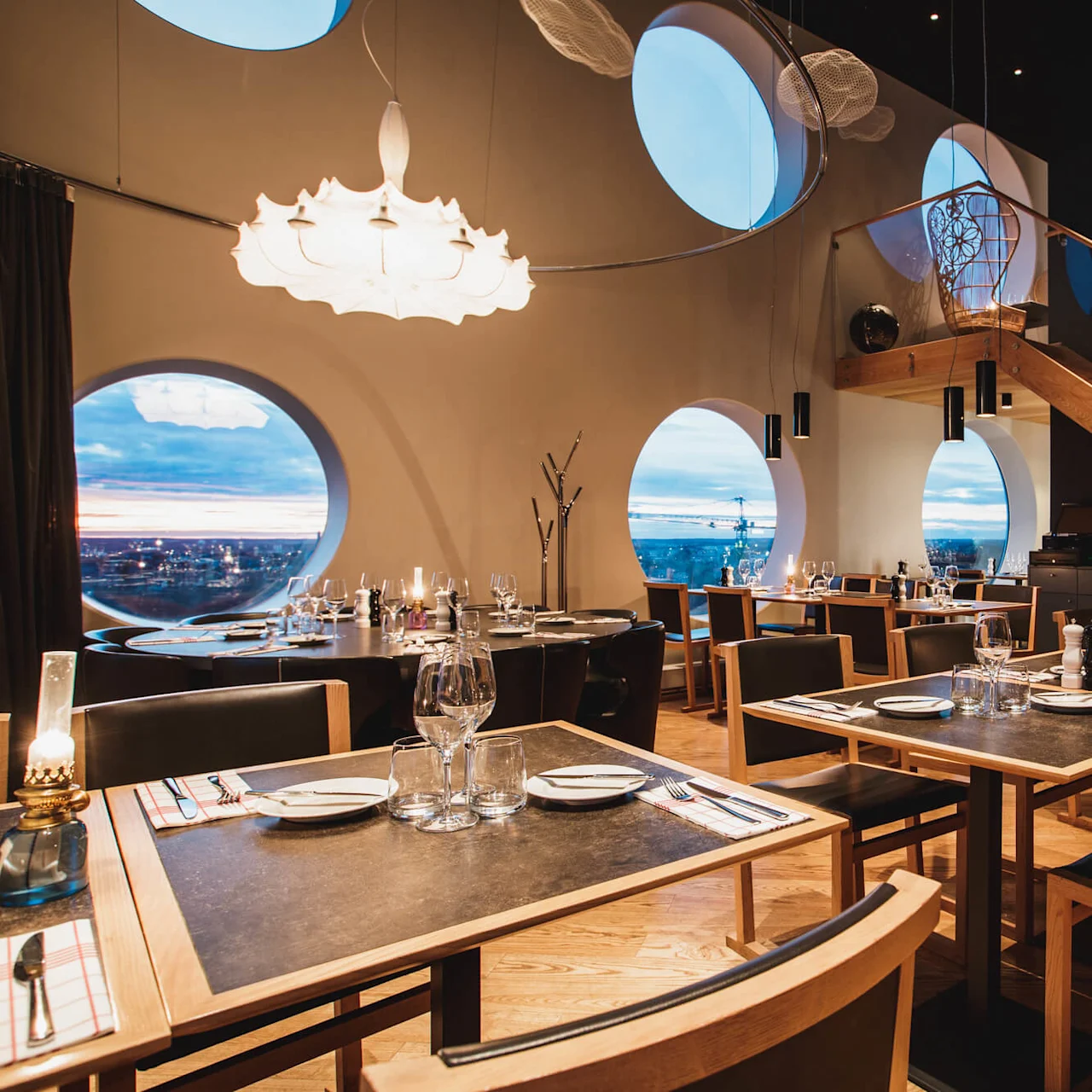 Elegante spiseborde dækket med glas og bestik i en moderne restaurant. Runde vinduer giver udsigt over byens skyline ved solnedgang. En trætrappe fører til en øvre etage.