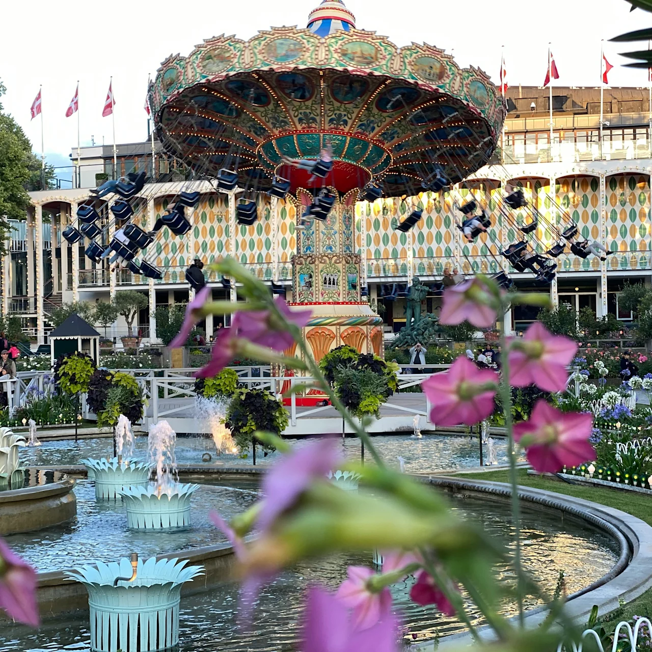 A vibrant amusement park scene with a swing carousel in motion, surrounded by fountains, lush greenery, and colorful flowers.