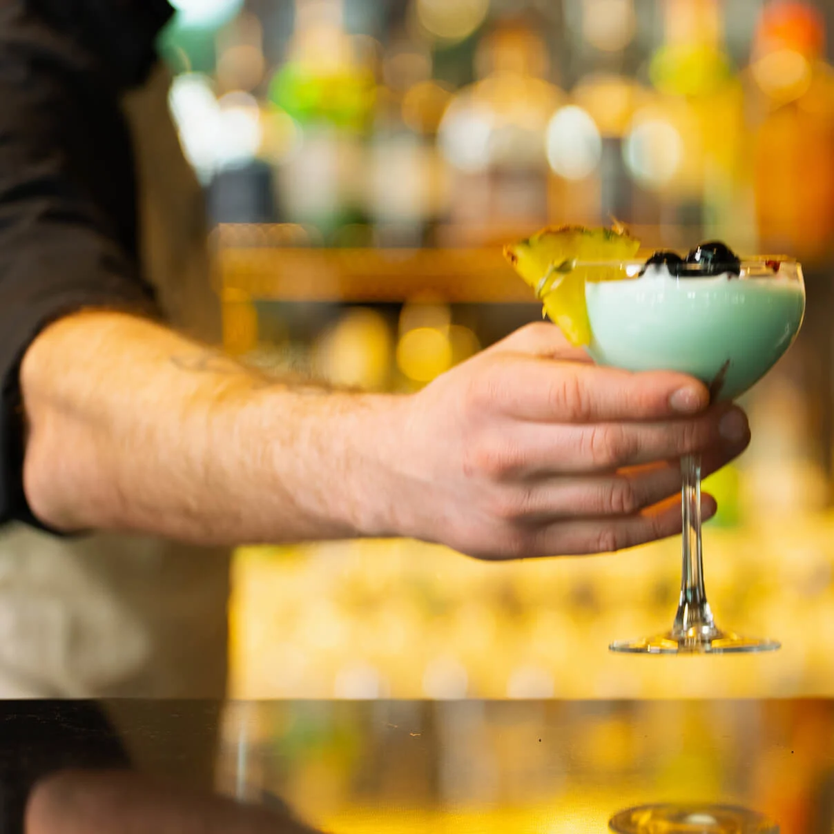En hand håller ett cocktailglas med fruktgarnering, mot en suddig bakgrund av en ljust upplyst bar med olika flaskor.