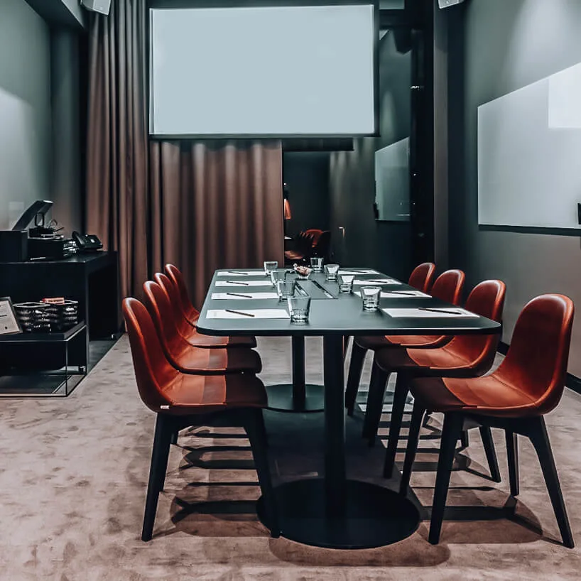 Meeting room with a long table set with notepads and glasses. Eight brown chairs surround the table. Artwork, whiteboard, and shelves complete the modern, minimalistic setting.
