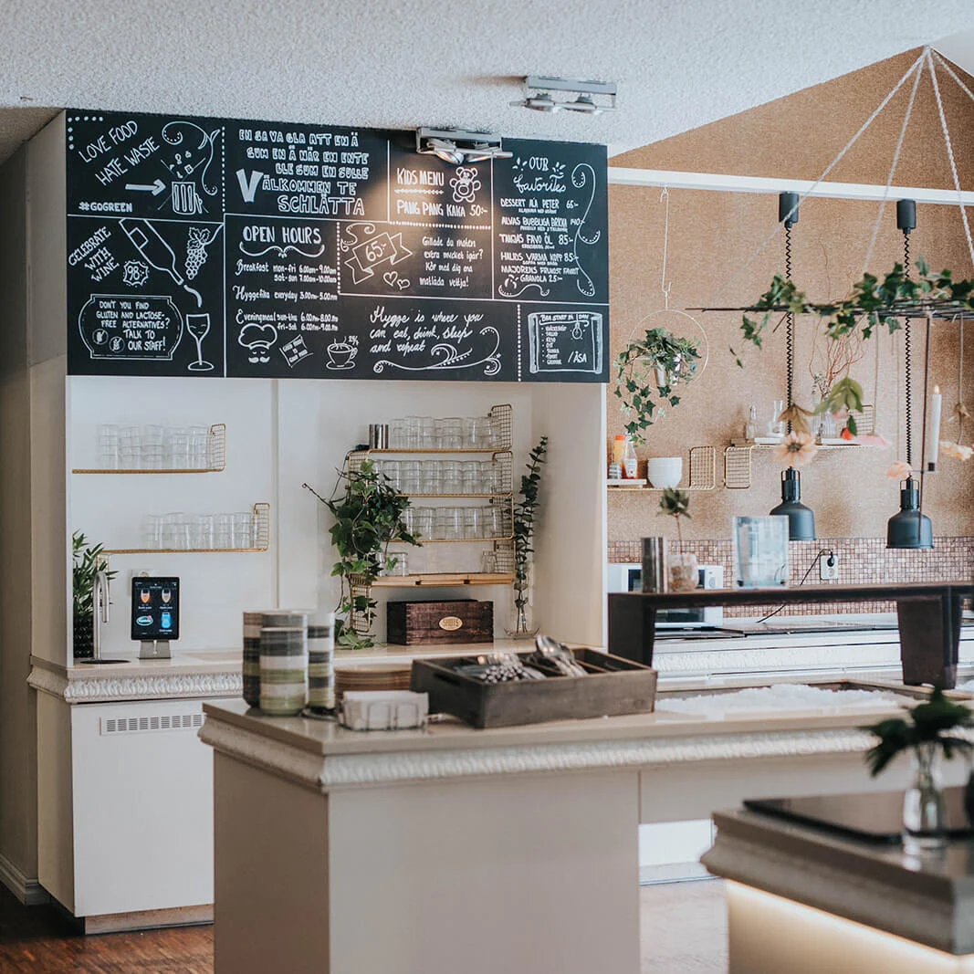 En hyggelig caféindretning med en stor kridttavle-menu, planter og en disk uden synlige mennesker.