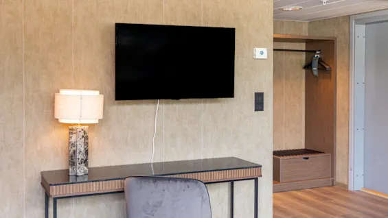 TV monterad på en beige vägg, ovanför ett skrivbord med en lampa och stol; bredvid finns en öppen garderob med galgar. Rumsmiljö med trägolv.