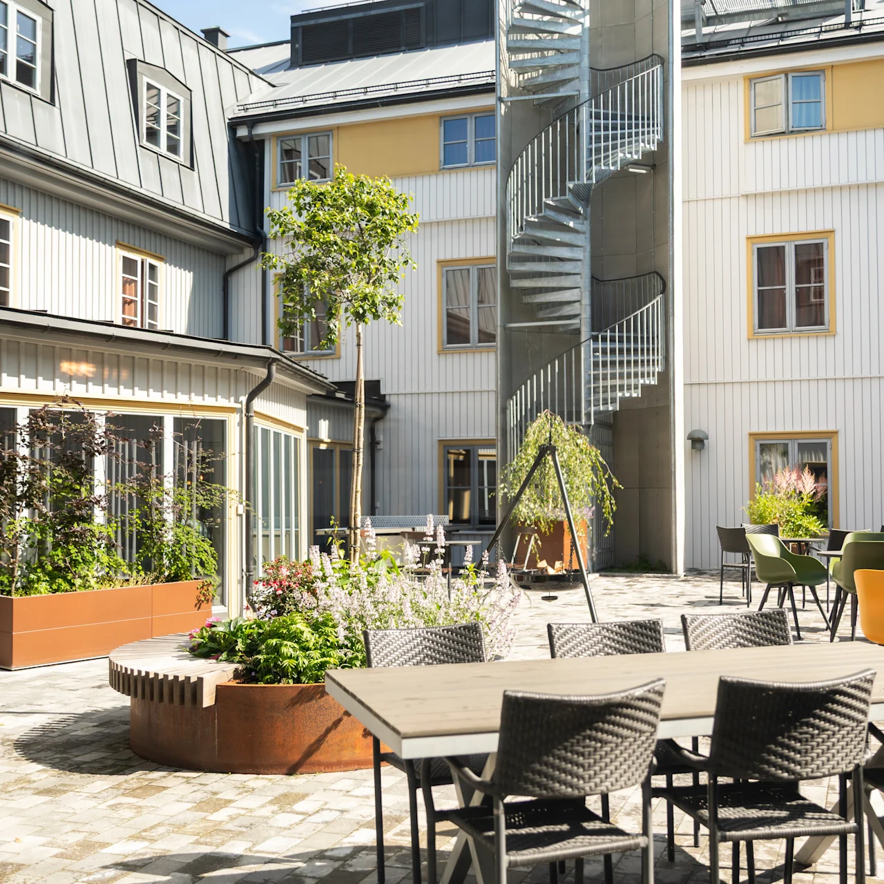 Udendørs gårdhave med spisebord og stole på stenlagt terrasse. Vindeltrappe fører op ad bygningens facade. Plantekasser og små træer pryder området.