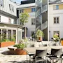 Udendørs gårdhave med spisebord og stole på stenlagt terrasse. Vindeltrappe fører op ad bygningens facade. Plantekasser og små træer pryder området.