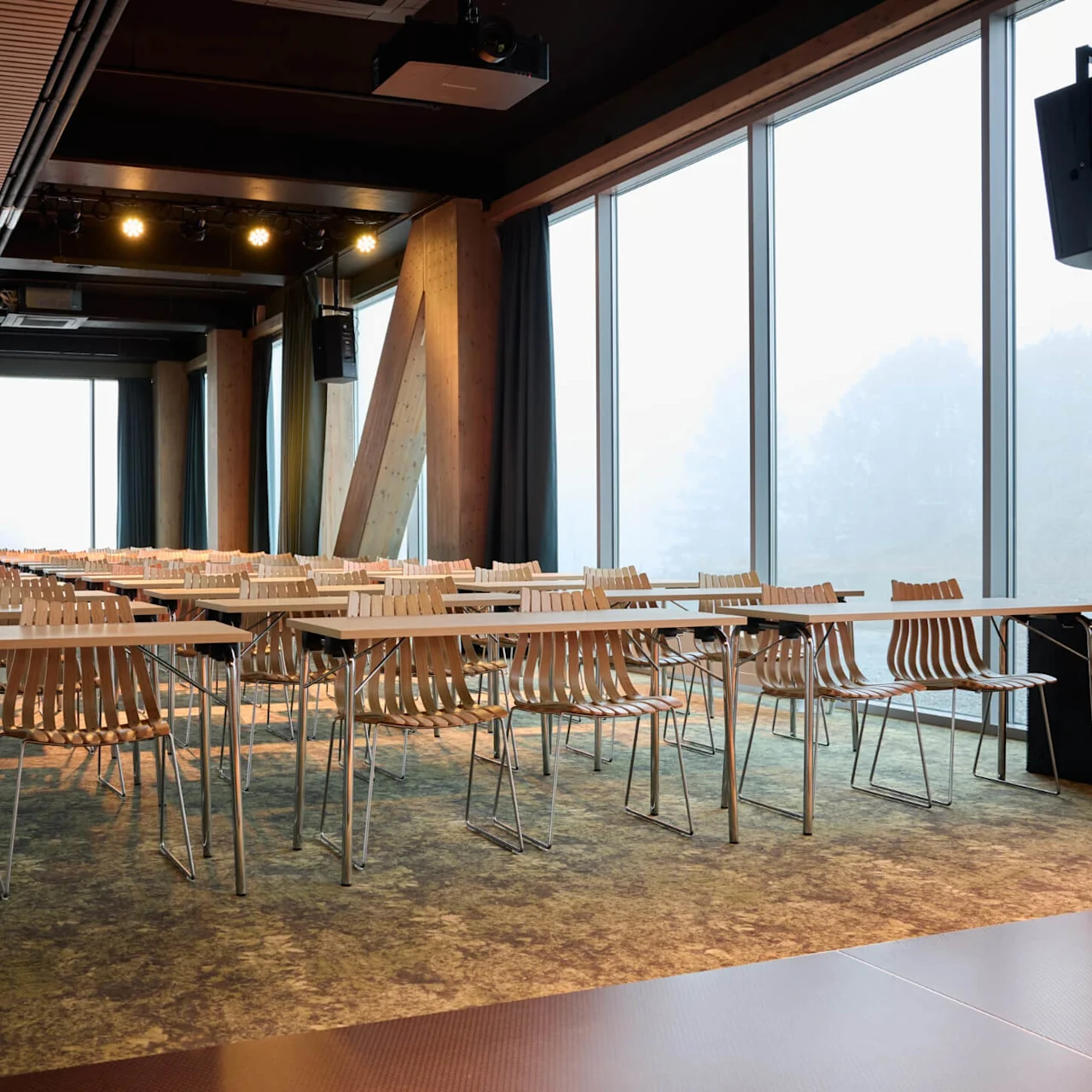 Wooden chairs and rectangular tables arranged in rows are in an empty conference room. Large windows line one side, allowing natural light to fill the space.