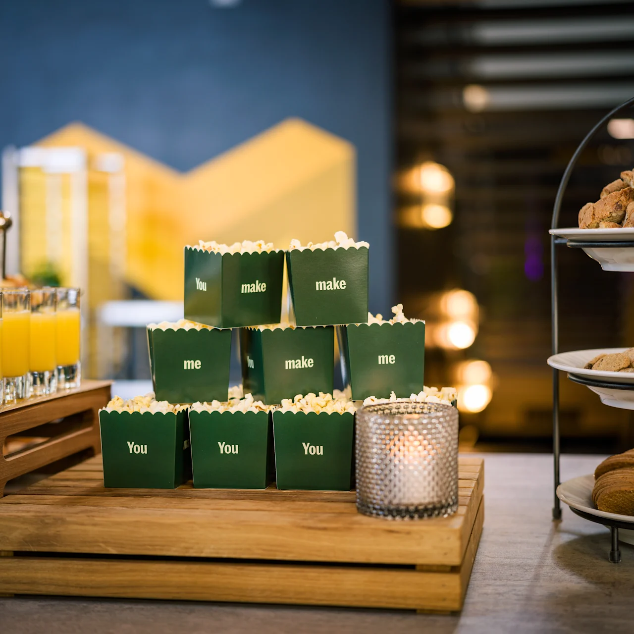 A stack of green popcorn boxes labeled "You make me" sits on a wooden tray; adjacent are orange juice shots and a tiered tray with cookies and biscotti.