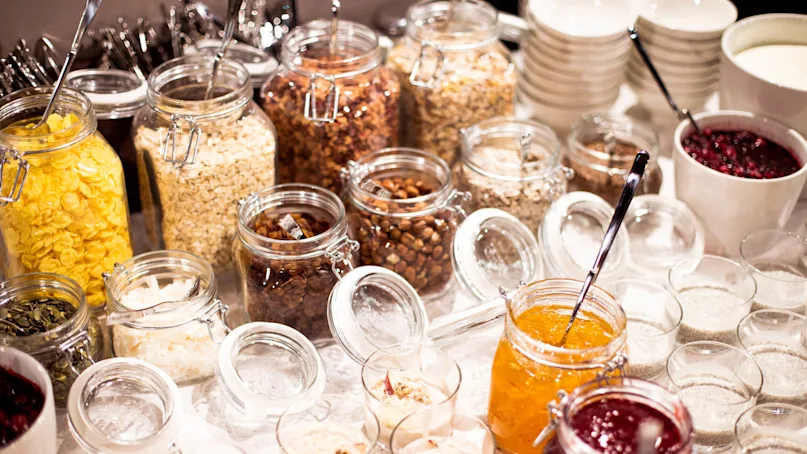 Various jars and bowls filled with breakfast items like cereals and jams are displayed on a table.