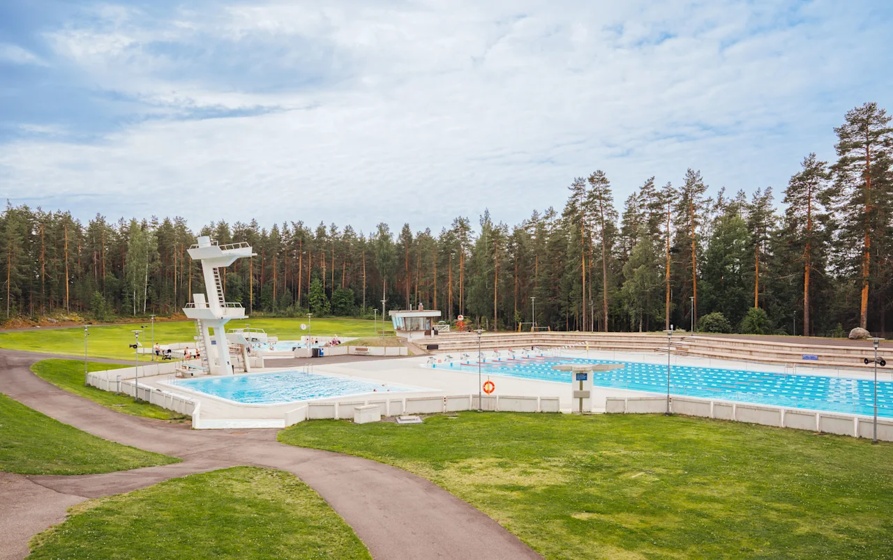 Yleisöuimala hyppytorneilla, jota ympäröi vihreä ruoho ja korkeat puut osittain pilvisellä taivaalla. Polut johtavat allasalueelle. Tekstiä ei ole.