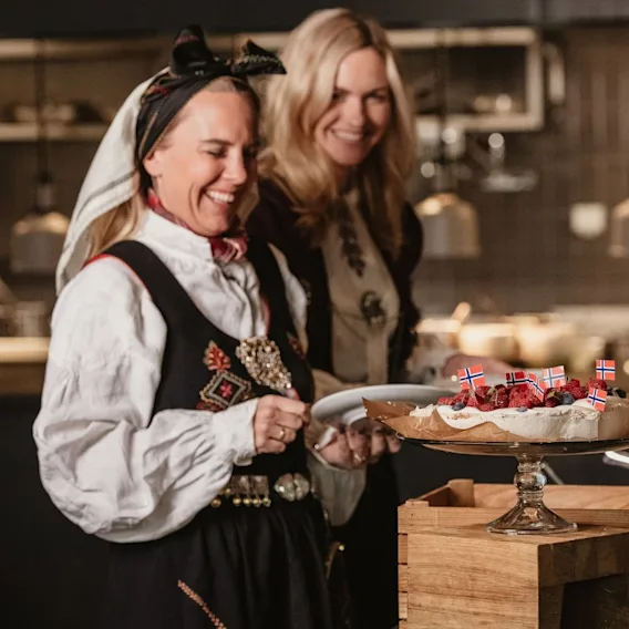 To smilende kvinner i bunad som serverer en kake pyntet med bær og små norske flagg.