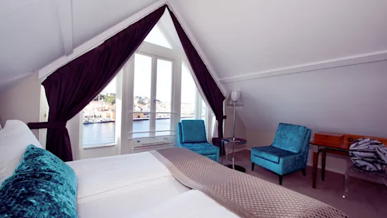 A neat hotel room with a bay window overlooking a scenic waterfront, velvet chairs, and plush bedding against a calm, neutral decor.