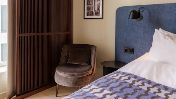 A cozy hotel room with a plush chair, an inviting bed with a blue headboard, and tasteful wall art, conveying a sense of warmth and comfort.