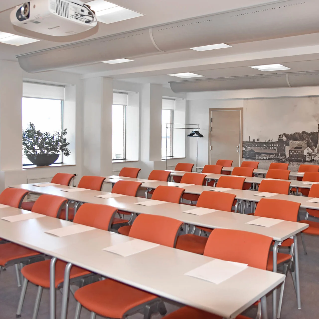 Classroom with rows of orange chairs and white desks holding paper sheets; overhead projector mounted on the ceiling, large mural of historic buildings on the back wall.