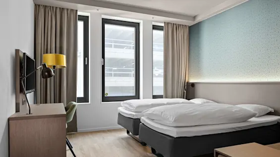 Two neatly made beds stand side by side in a modern hotel room, featuring light wooden floors, a patterned blue accent wall, and large windows with beige curtains.