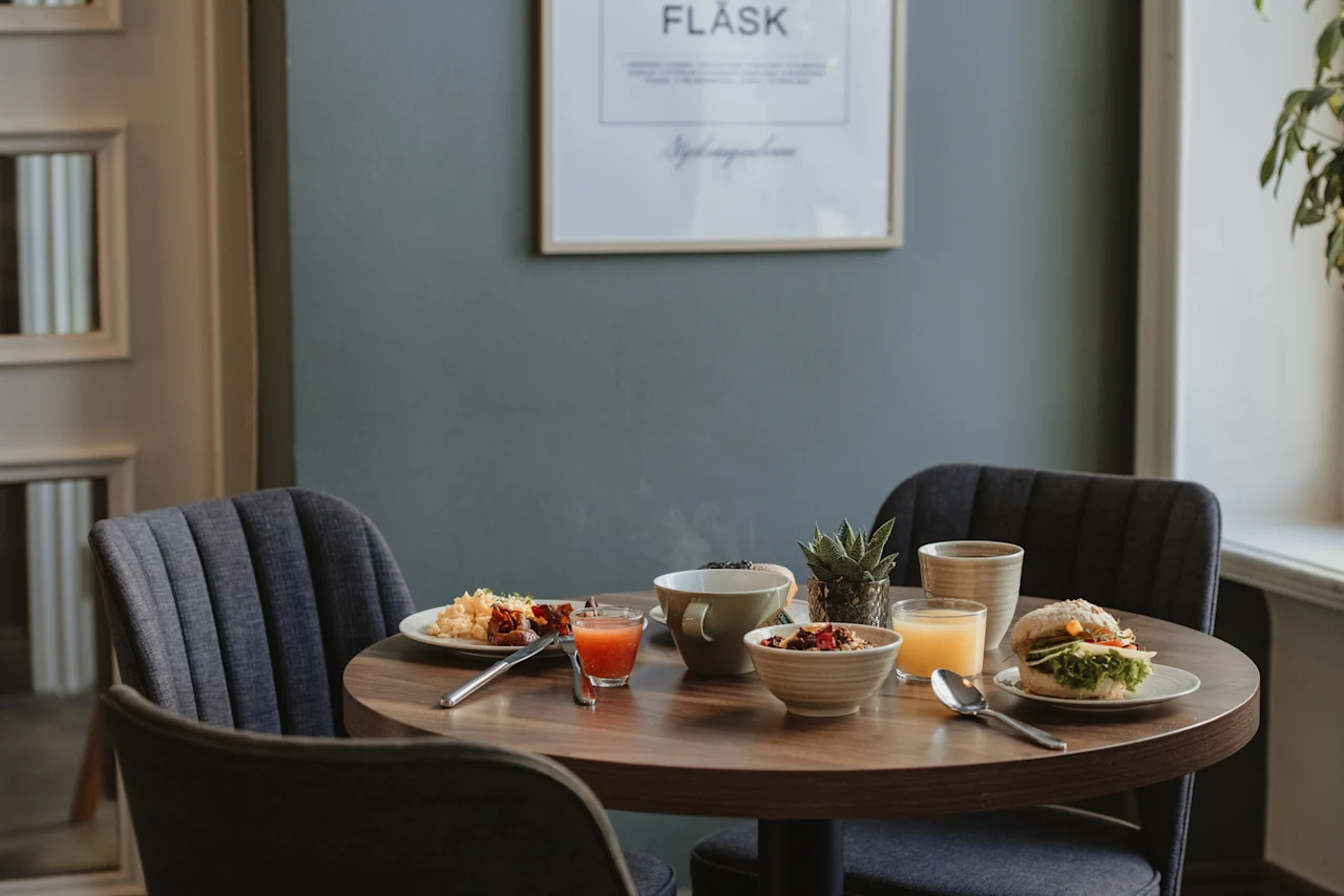 Ett hotellfrukostbord dukat för två med olika rätter, juicer och en varm dryck. En inramad bild med texten FLASK hänger på väggen.