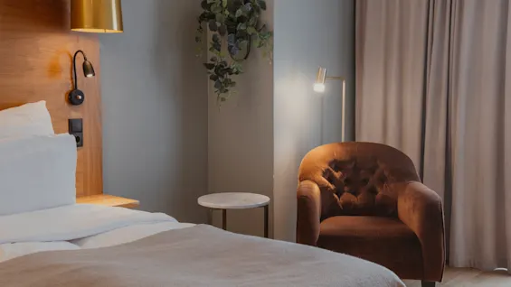 Et hyggeligt hotelværelse med en komfortabel seng, en blød lænestol og varm belysning, der indbyder til afslapning og hvile.