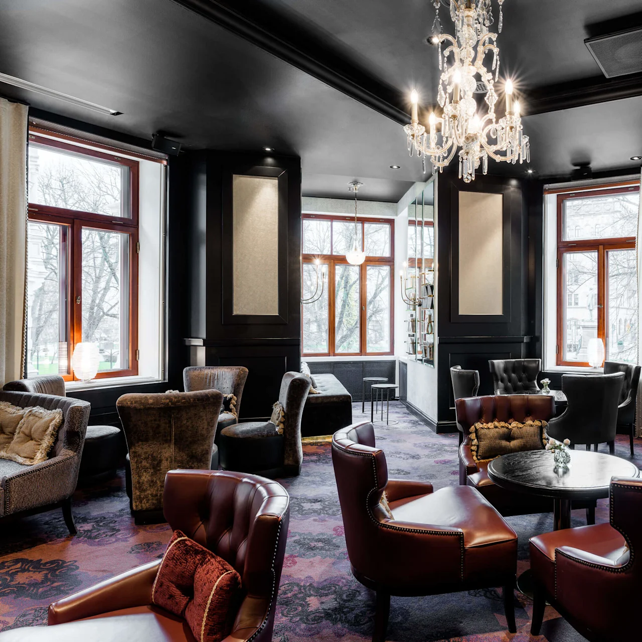 An elegant lounge with leather chairs, plush sofas, a crystal chandelier, and large windows providing natural light at Hotel Kämp in Helsinki.