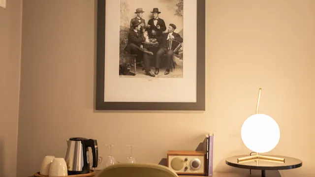 Et innrammet svart-hvitt bilde henger over et skrivebord. Skrivebordet har en lampe, radio, bøker, glass, kopper og en vannkoker. Innstillingen er et mykt opplyst, minimalistisk rom.