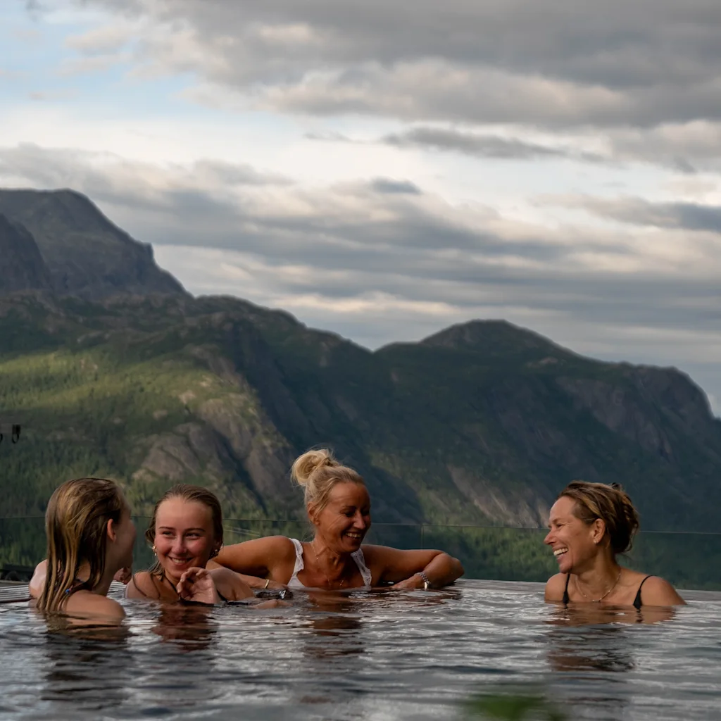 Fire personer slapper af i en infinity pool, smiler og snakker mod en baggrund af frodig, bjergrig natur under en skyet himmel.