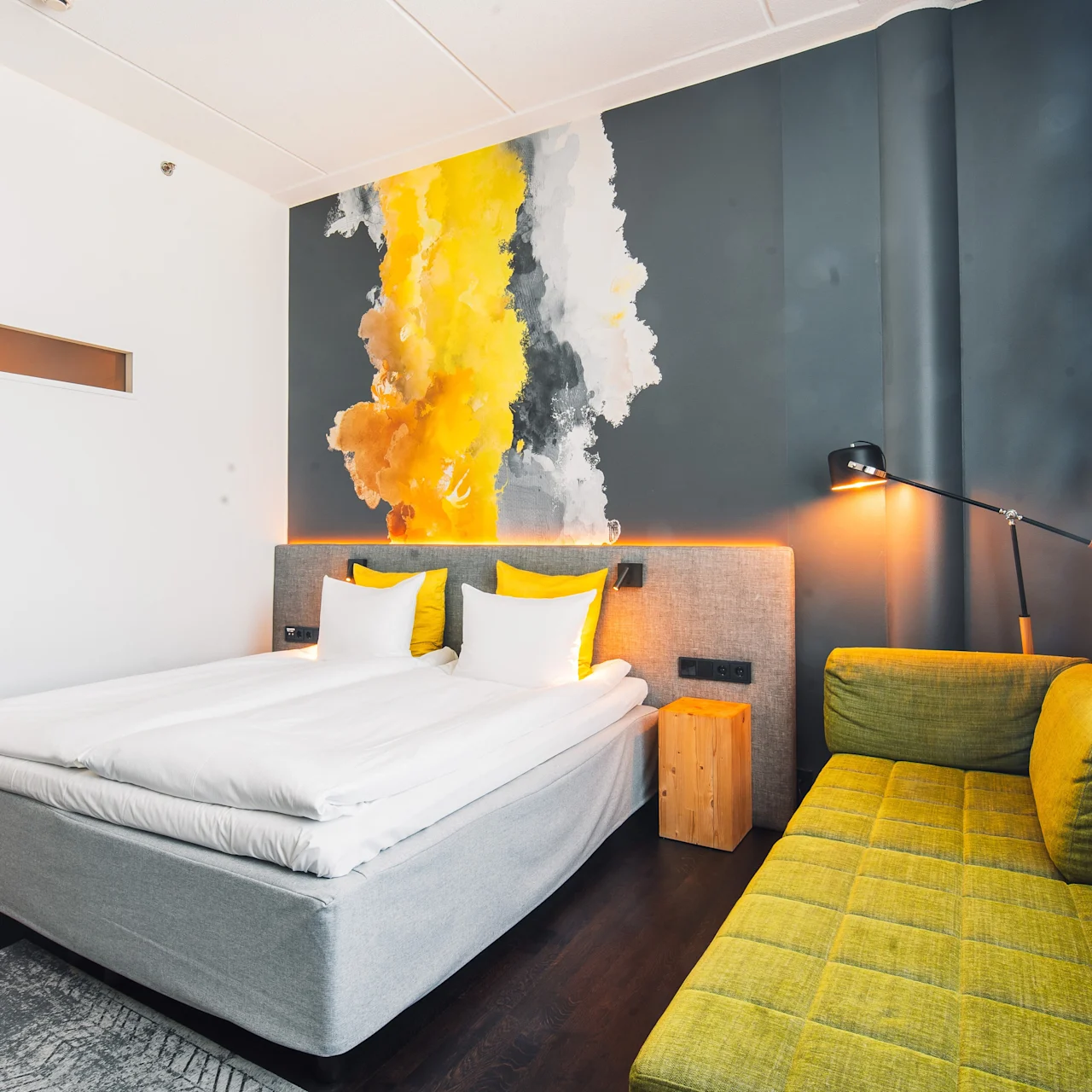 Bed resting beside a yellow, gray, and white abstract wall mural, with green sofa and floor lamp in a hotel room. Light enters through a large window with sheer curtains.