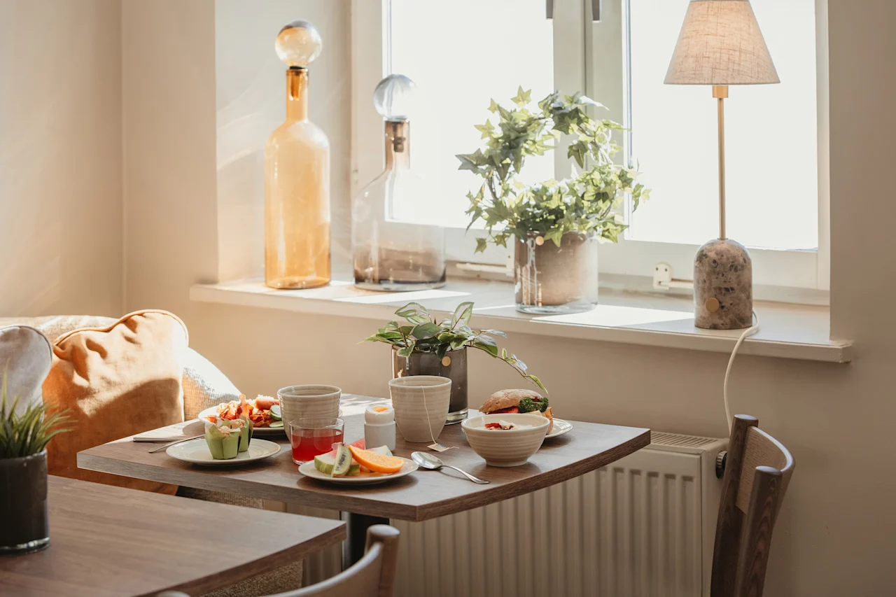 A cozy breakfast spread on a table by a sunlit window, with fresh fruit, a sandwich, and drinks.