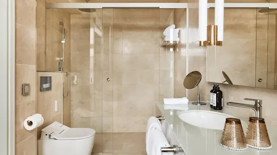 Et moderne badeværelse med glasbruser, beige fliser og et slankt toilet. En bordplade holder et rundt spejl, sæber og håndklæder, med omgivende belysning.