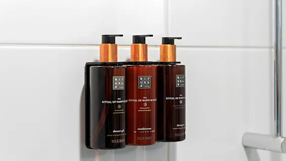Three brown bottles with pump tops rest on a bath ledge, labeled shower gel, conditioner, and shampoo, in a white-tiled bathroom.