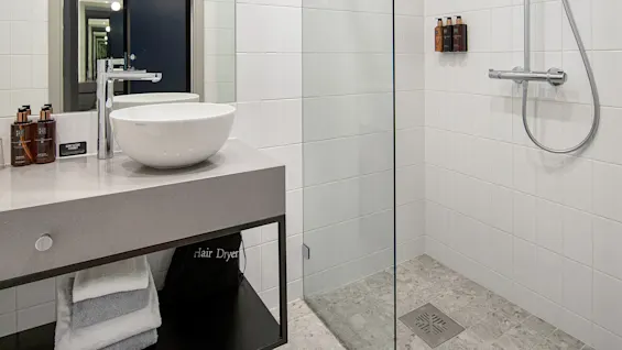 Sink sits atop a countertop beside toiletries and a mirror; glass shower enclosure and tiled walls surround. Towels lie below. Text: "Hair Dryer."