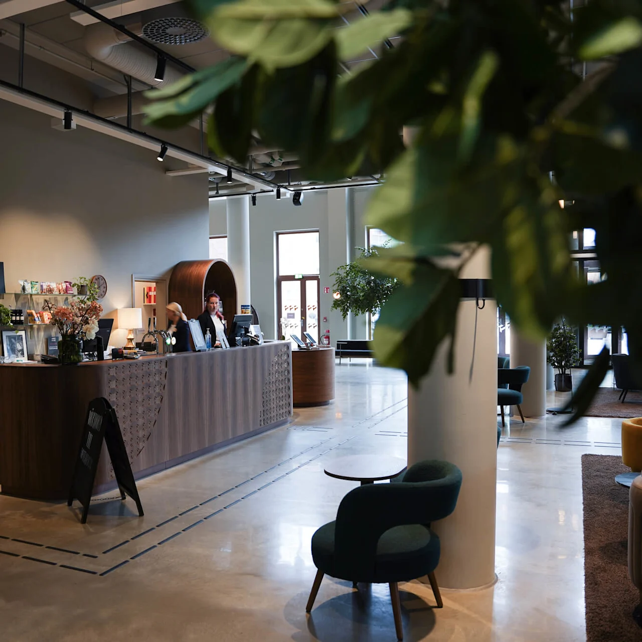 Modern lobby with a reception desk, staff, and seating areas, framed by indoor plants; a bright, inviting atmosphere.