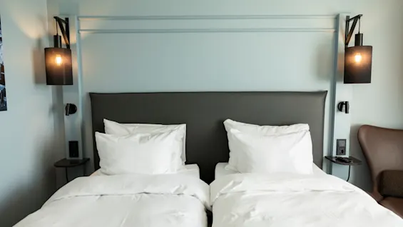 To enkeltsenger med hvitt sengetøy og puter, en mørk sengegavl og veggmonterte lamper på et hotellrom.