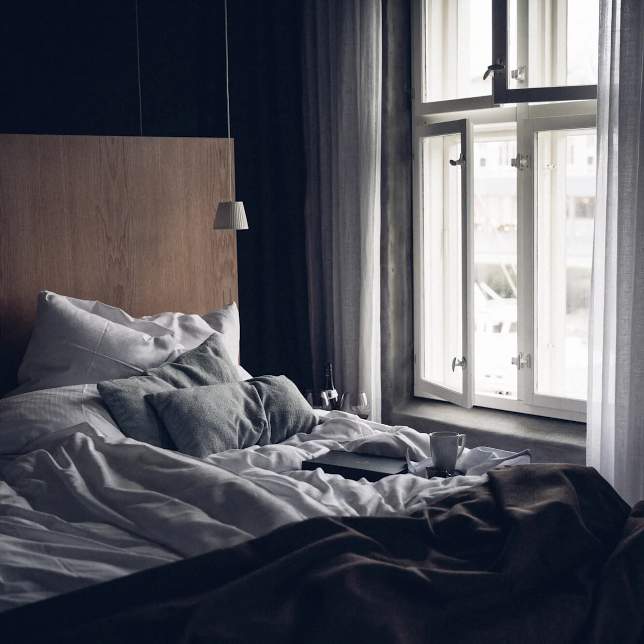 A neatly made bed with white and gray pillows, brown blanket, and open windows; a mug and closed book lie atop. The room appears calm and softly lit.