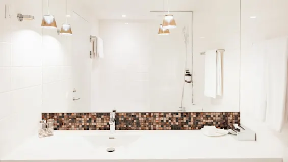 Et moderne hvidt badeværelse har et stort spejl og mosaikbagvæg, med hængende lamper over vasken. Håndklædestænger og brusehoved er synlige, hvilket skaber en ren og minimalistisk atmosfære.