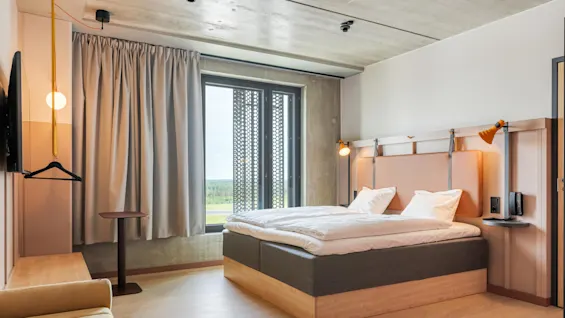 Et moderne hotelværelse har en pænt redt seng med hvidt linned, blød belysning og minimalistiske møbler, inklusiv en sofa, sidebord og et stort vindue med gardiner.