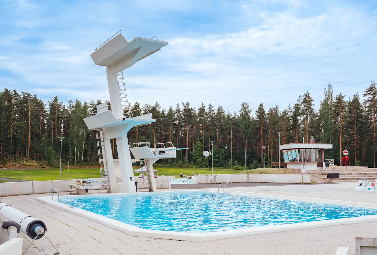 Suuri ulkouima-allas, jossa on useita uimahyppyalustoja, ympäröity korkealla metsällä pilvisen taivaan alla. Taustalla näkyy pieni rakennus.