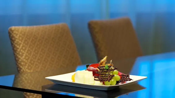 A plate with assorted cheeses, fruits, and chocolate garnish rests on a glass table with blurred chairs and blue backdrop.