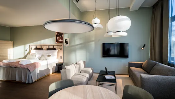 A modern hotel room features a neatly made double bed, a round table with chairs, and a cozy seating area with a sofa facing a mounted television, under stylish ceiling lamps.